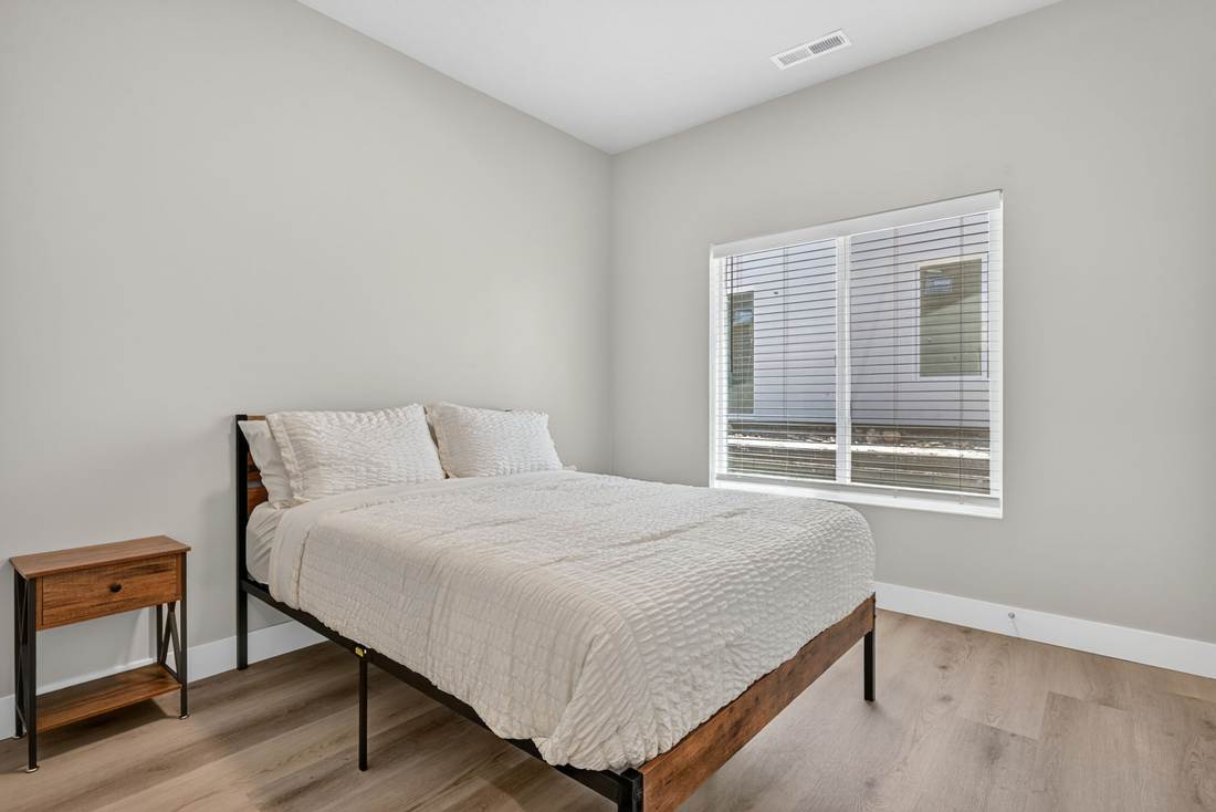 Bedroom with a queen bed, wood and metal frame, nightstand, and a wide blinded window.