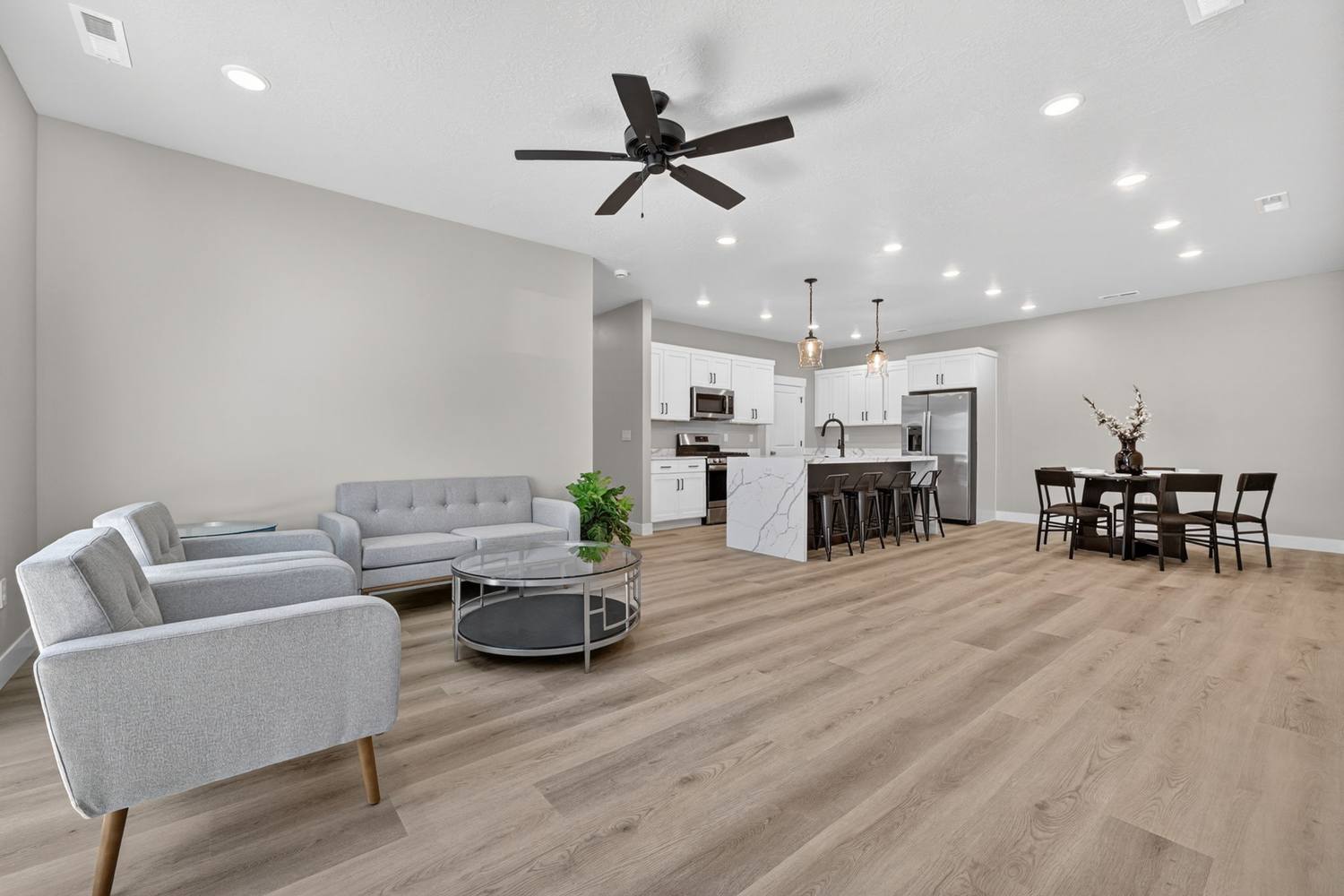 Open great room with grey upholstered seating, glass coffee table, ceiling fan, and sightline through to the kitchen and dining area.