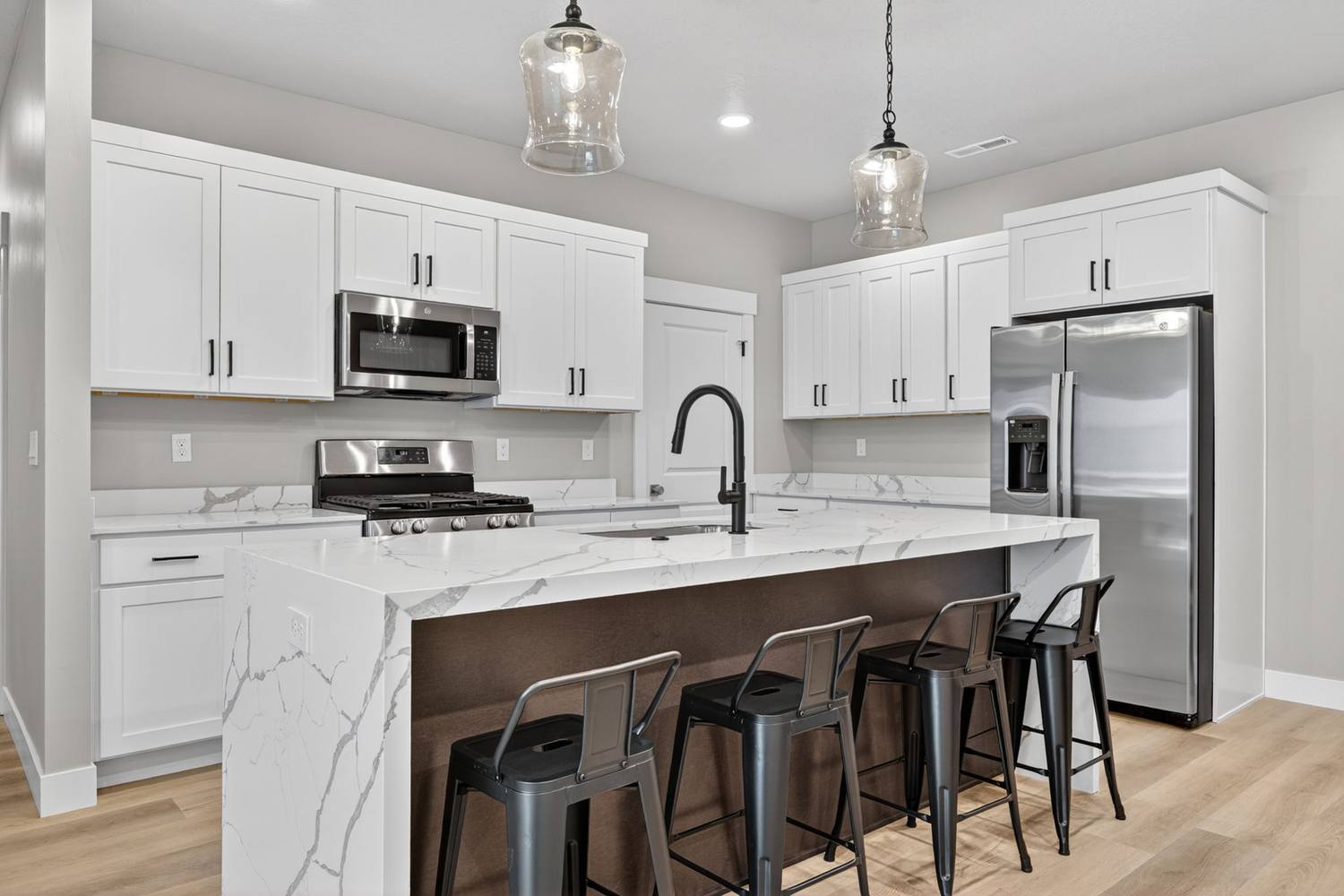 The kitchen island in waterfall-edge quartz with metal barstools, black gooseneck faucet, white shaker cabinetry and a gas range behind.
