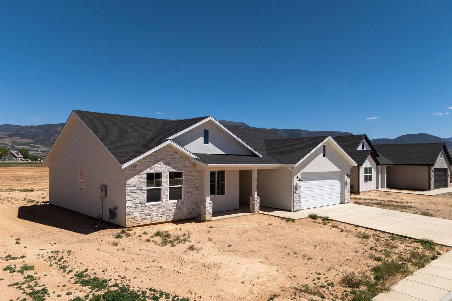 5174 Sagewood, a single-level stucco and stone custom home with a two-car garage, mountains rising behind it.