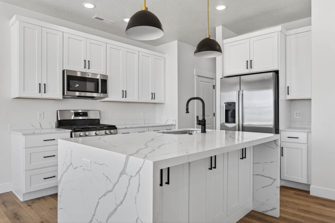 Full-slab waterfall quartz island with white shaker cabinetry, stainless gas range and refrigerator behind.